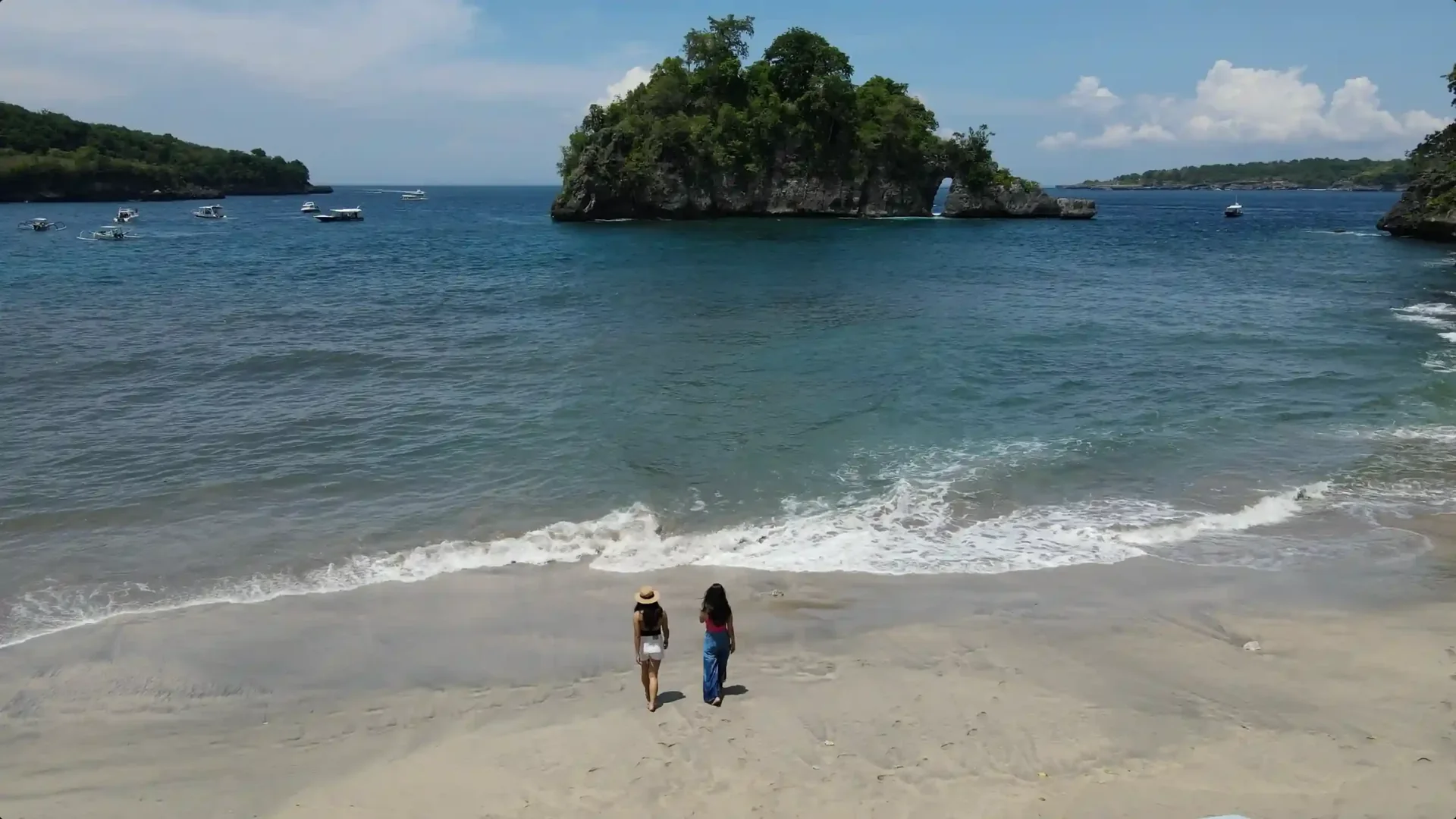Two people walking an empty sand beach with day boats moored offshore